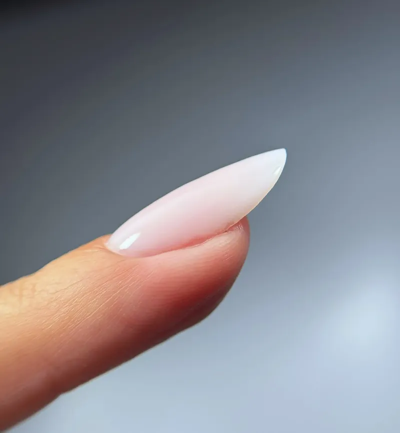 Ongle amande long, rose lacté, vu de profil sur fond gris.